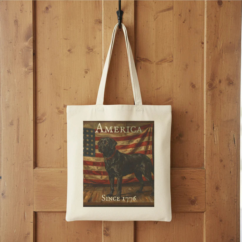 A cream canvas tote bag hanging from a black hook on a rustic wooden wall. The bag features a printed graphic of a black lab standing in front of an American flag, with the text 'AMERICA' at the top and 'SINCE 1776' at the bottom.