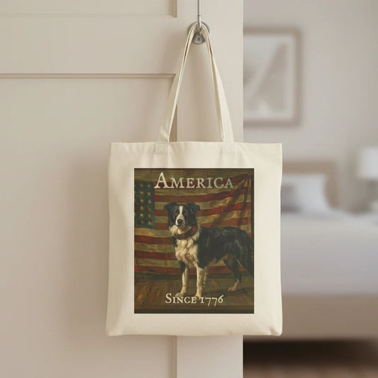 An off-white canvas tote bag hanging from a metal hook on a white door in a softly blurred bedroom setting. The bag features a printed graphic of a Border Collie standing in front of an American flag, with the text 'AMERICA' at the top and 'SINCE 1776' at the bottom.