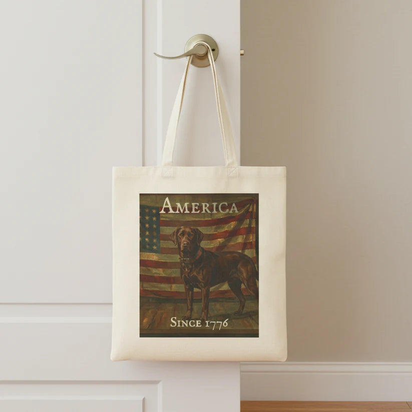 An off-white canvas tote bag hanging from a brass doorknob on a white door. The bag features a vintage-style graphic print of a Chocolate Lab standing in front of an American flag, with the text 'AMERICA' at the top and 'SINCE 1776' at the bottom.