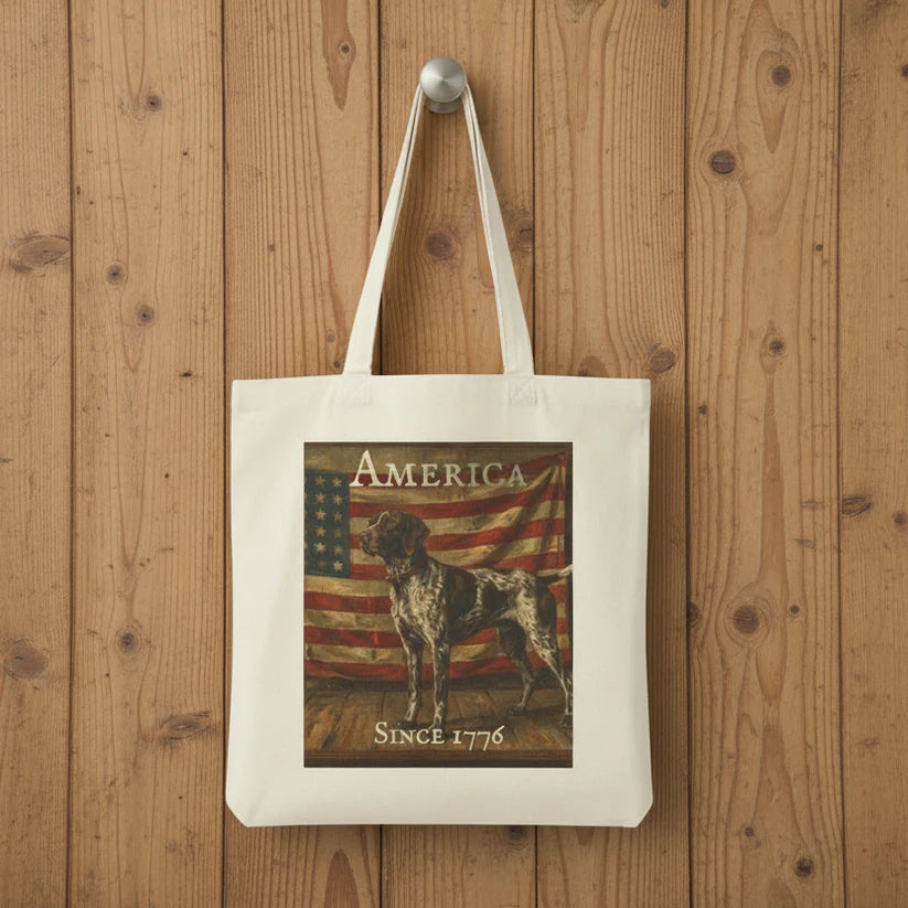 A white canvas tote bag with long handles, featuring a printed graphic of a German Shorthaired Pointer dog in front of an American flag with the text 'AMERICA' and 'SINCE 1776', hanging on a silver hook against a rustic wooden plank background.