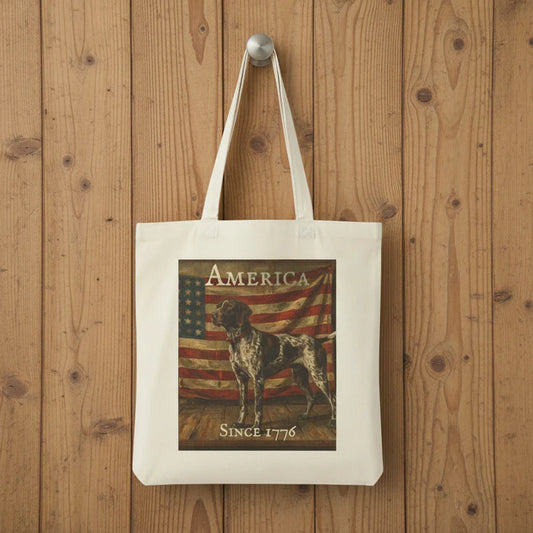 A white canvas tote bag with long handles, featuring a printed graphic of a German Shorthaired Pointer dog in front of an American flag with the text 'AMERICA' and 'SINCE 1776', hanging on a silver hook against a rustic wooden plank background.