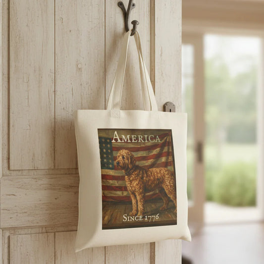 An off-white canvas tote bag hanging from a black hook on a light wood-paneled door. The bag features a printed graphic of a goldendoodle dog in front of an American flag, with the text 'AMERICA' at the top and 'SINCE 1776' at the bottom.