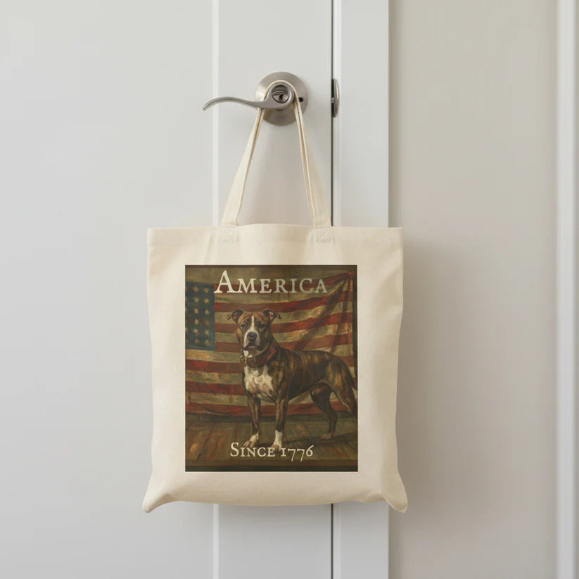 A cream canvas tote bag hanging from a silver doorknob on a white paneled door. The bag features a vintage-style graphic print of a brindle pitbull standing in front of an American flag, with the text 'AMERICA' at the top and 'SINCE 1776' at the bottom.