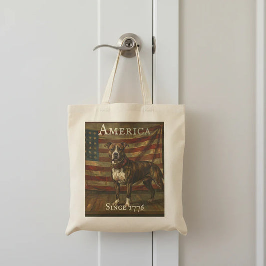 A cream canvas tote bag hanging from a silver doorknob on a white paneled door. The bag features a vintage-style graphic print of a brindle pitbull standing in front of an American flag, with the text 'AMERICA' at the top and 'SINCE 1776' at the bottom.