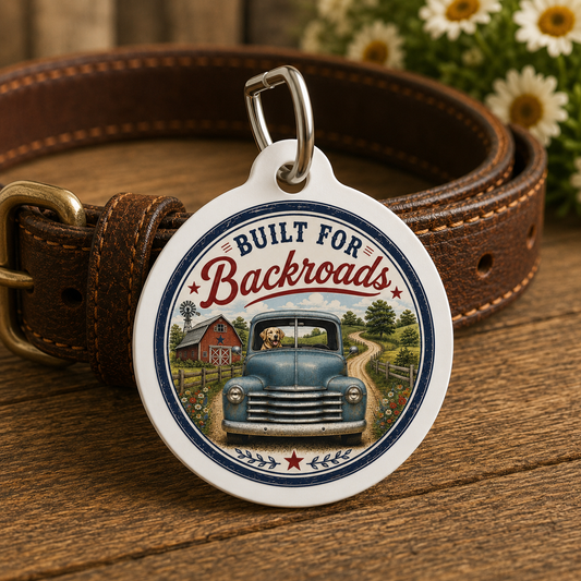 A round pet ID tag with a white border and a colorful illustration of a blue truck on a country road, attached to a brown leather collar. The collar is placed on a wooden surface with blurred daisies in the background.