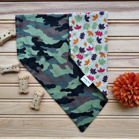 A reversible dog bandana with a camo pattern on one side and fall leaves on the other, displayed on a wooden surface with cookies and a pumpkin decoration nearby.