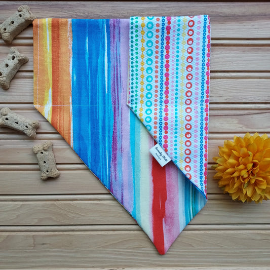 A reversible dog bandana with a colorful striped and dotted pattern, displayed on a wooden surface with dog treats and a yellow flower nearby.