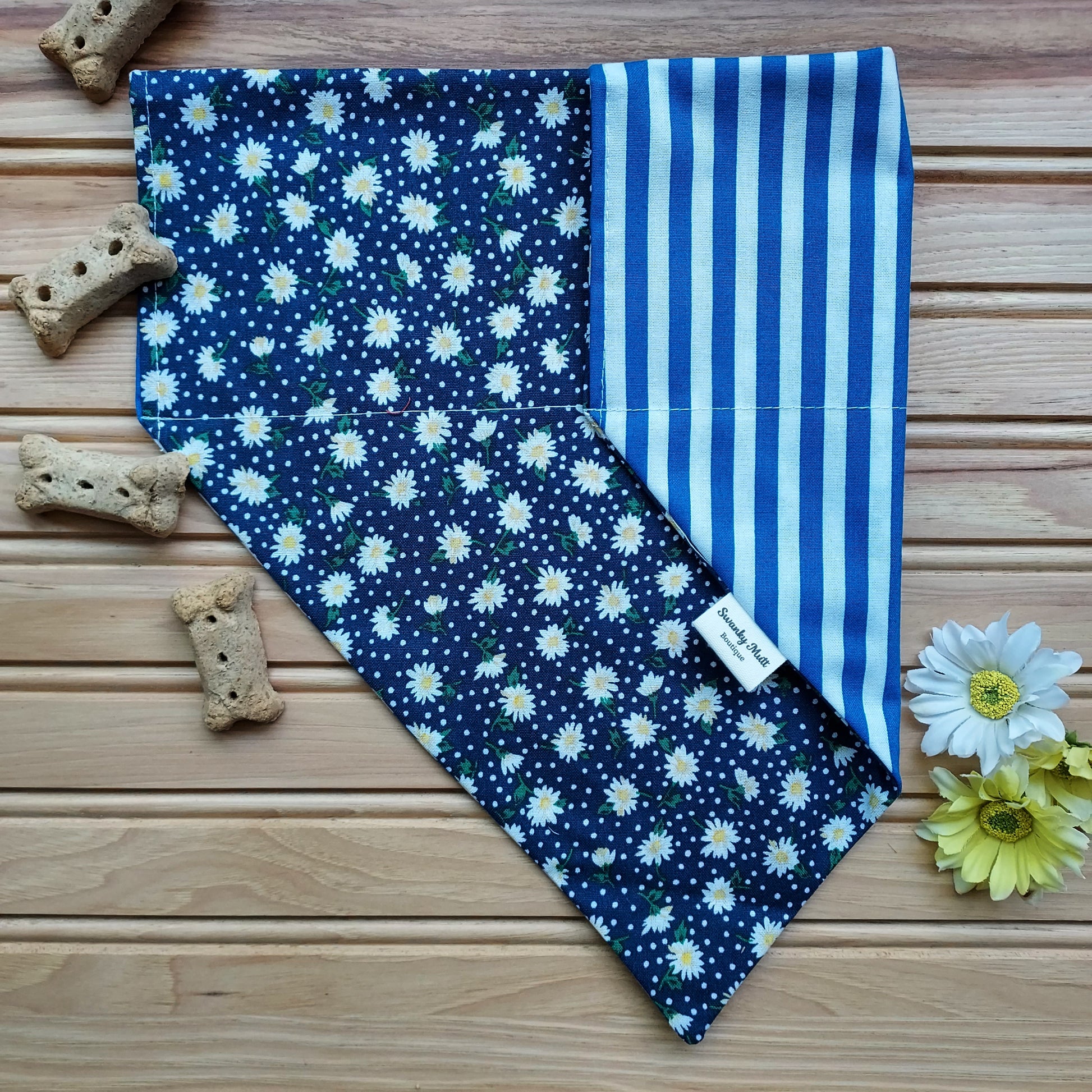 A reversible dog bandana with a white daisy pattern on one side and blue and white stripes on the other, displayed on a wooden surface with dog biscuits and flowers around it.