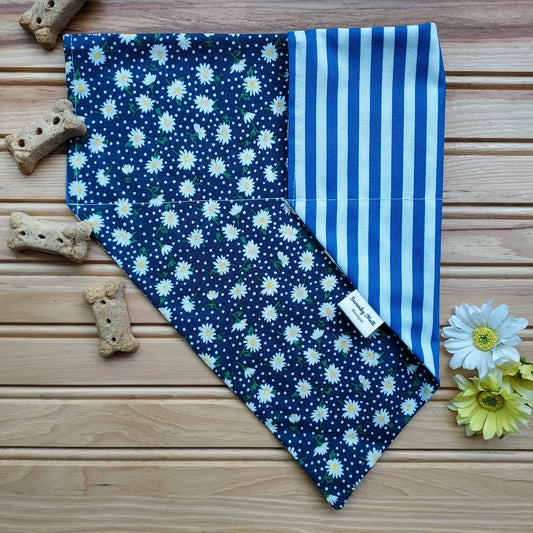 A reversible dog bandana with a white daisy pattern on one side and blue and white stripes on the other, displayed on a wooden surface with dog biscuits and flowers around it.