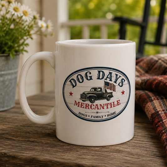 A white ceramic mug with a 'Dog Days Mercantile' vintage truck graphic, set on a rustic wooden table next to a plaid blanket and a potted daisy.