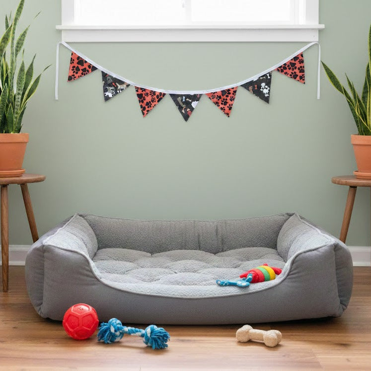 Gray pet bed with toys on a wooden floor, surrounded by potted plants and a decorative banner.