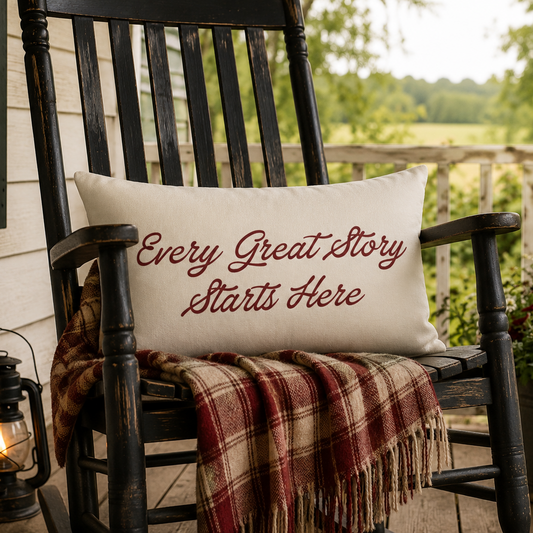 A white rectangular throw pillow with the phrase 'Every Great Story Starts Here' printed in dark red script, resting on a black rocking chair on a porch. A red, beige, and brown plaid blanket is draped over the chair, with a lit lantern on the left and a potted American flag and flowers on the right.