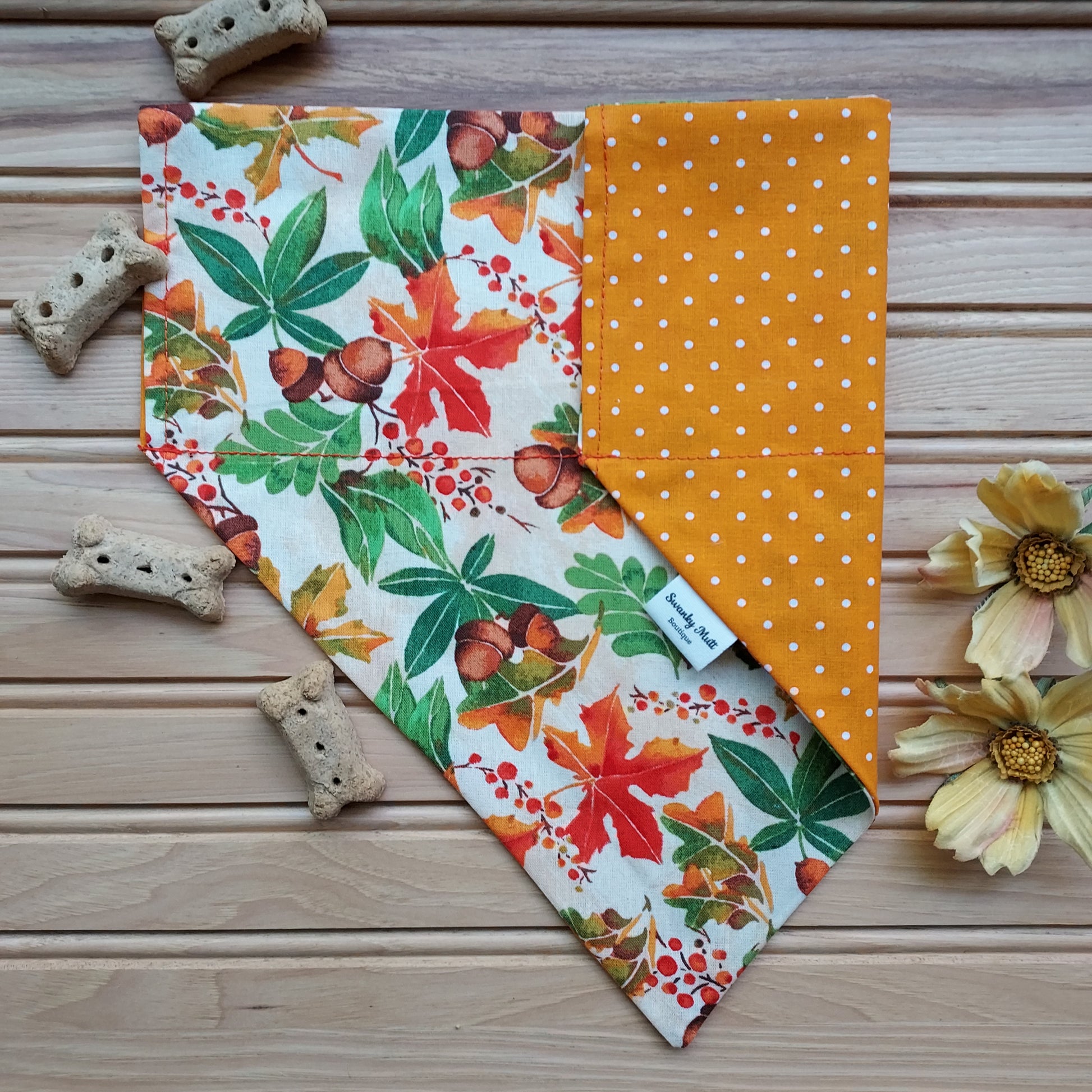 A reversible dog bandana with a fall leaves print on one side and orange dots on the other, displayed on a wooden surface with dog treats and flowers nearby.