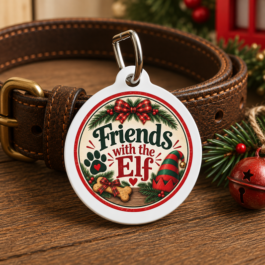 A round white pet ID tag with a silver clasp, featuring a festive Christmas design with the text 'Friends with the Elf' and holiday illustrations, shown attached to a brown leather dog collar on a wooden surface with holiday decorations.