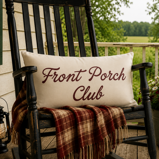 A cream rectangular throw pillow with 'Front Porch Club' embroidered in dark red script, resting on a black rocking chair on a porch. A red, brown, and beige plaid blanket is draped over the chair, with a lantern on the left and a potted flower arrangement with a small American flag on the right.