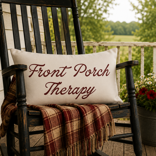A cream rectangular throw pillow with the phrase 'Front Porch Therapy' embroidered in dark red script, resting on a black rocking chair on a porch. A red, brown, and beige plaid blanket is draped over the chair, alongside a lit lantern, a potted red flower arrangement, and a small American flag, with a blurred green outdoor background.