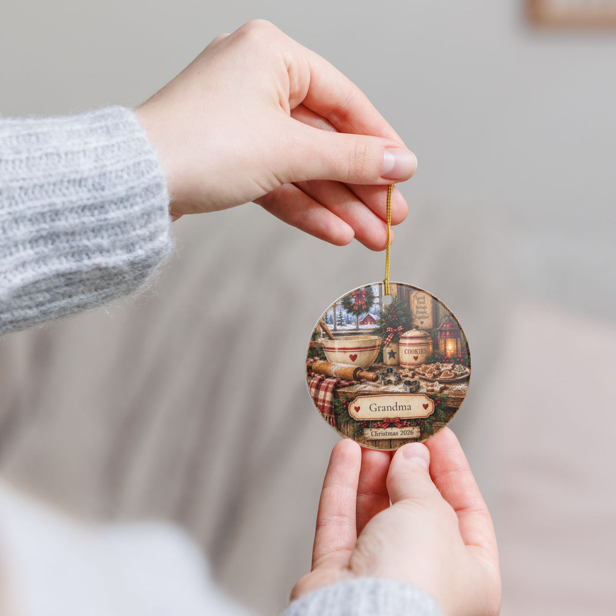 A person holds a round personalized Christmas ornament with a gold hanging string. The ornament features a rustic printed scene with barrels, a lantern, and the text 'Grandma' and 'Christmas 2026'.