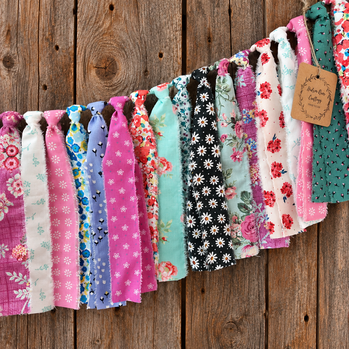 A fabric garland banner made of multiple fabric strips in assorted floral and patterned prints, hung against a rustic wooden plank background. A small paper tag is attached on the right side.