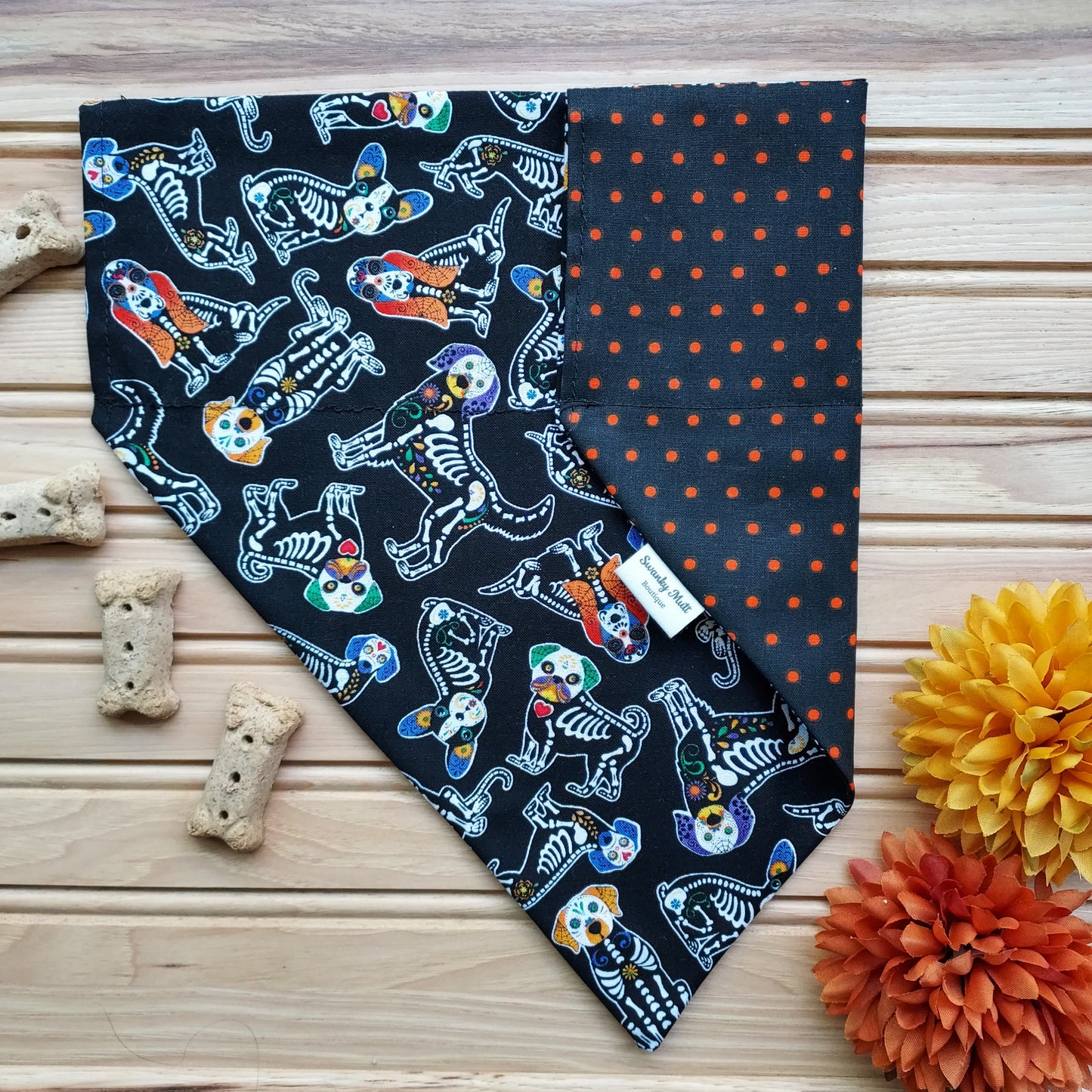 A reversible dog bandana with a black base featuring a colorful skeleton pattern and an orange base with a black dot pattern. The bandana is displayed on a wooden surface with a pumpkin and dog treats, and it appears to be made of a soft fabric.