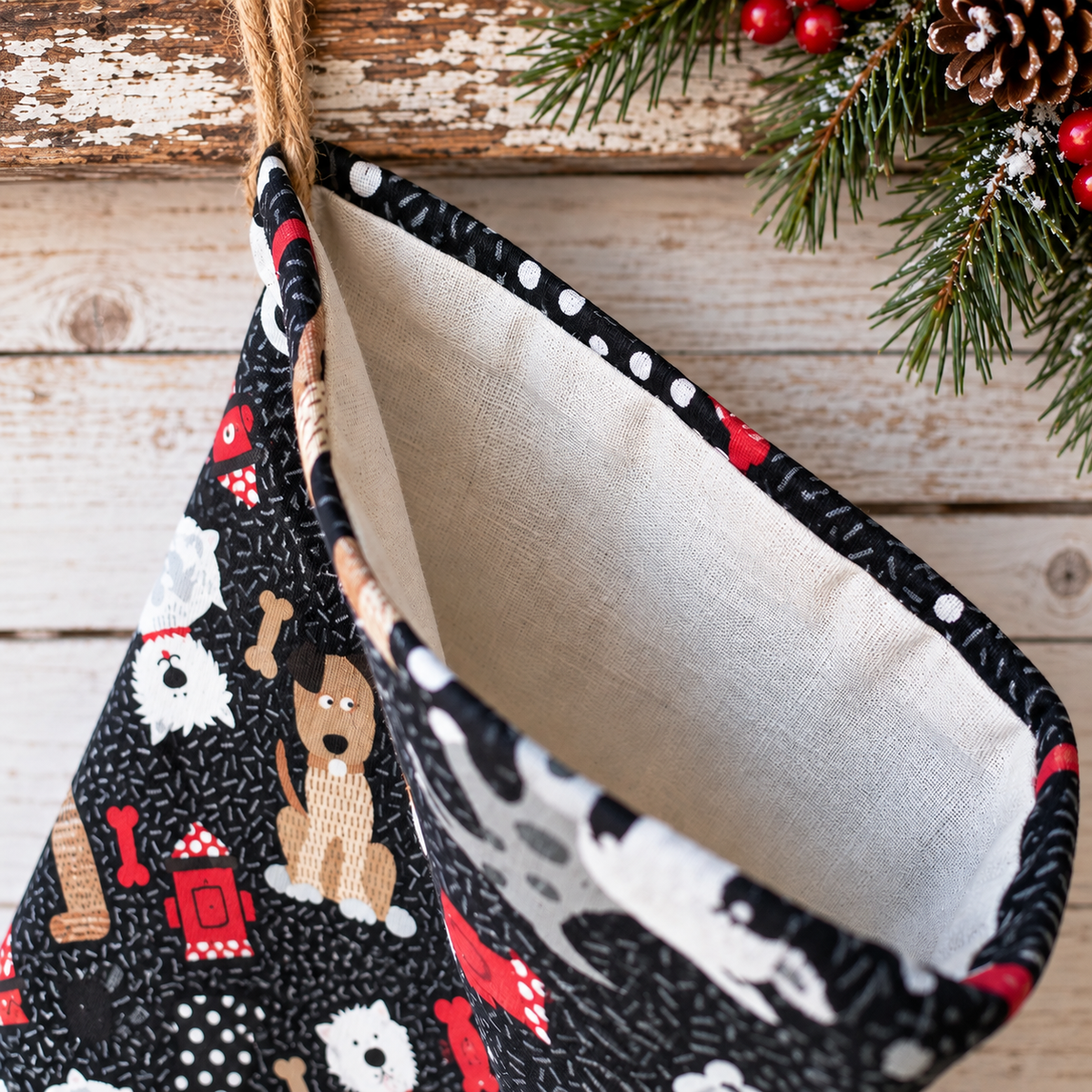 A close-up of a Christmas stocking with a black background fabric printed with various dog breeds, red ornaments, and white polka dots, hanging from a jute rope against a rustic wooden background with holiday greenery.