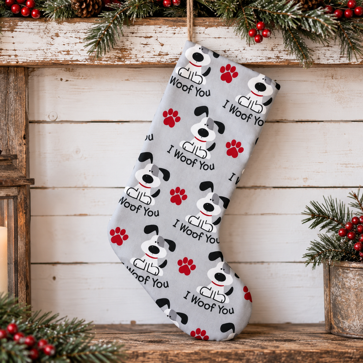 A gray Christmas stocking with a repeating pattern of black and white dog faces, red paw prints, and the text 'I Woof You', hanging against a rustic white wood plank wall with holiday greenery and red berries around it.