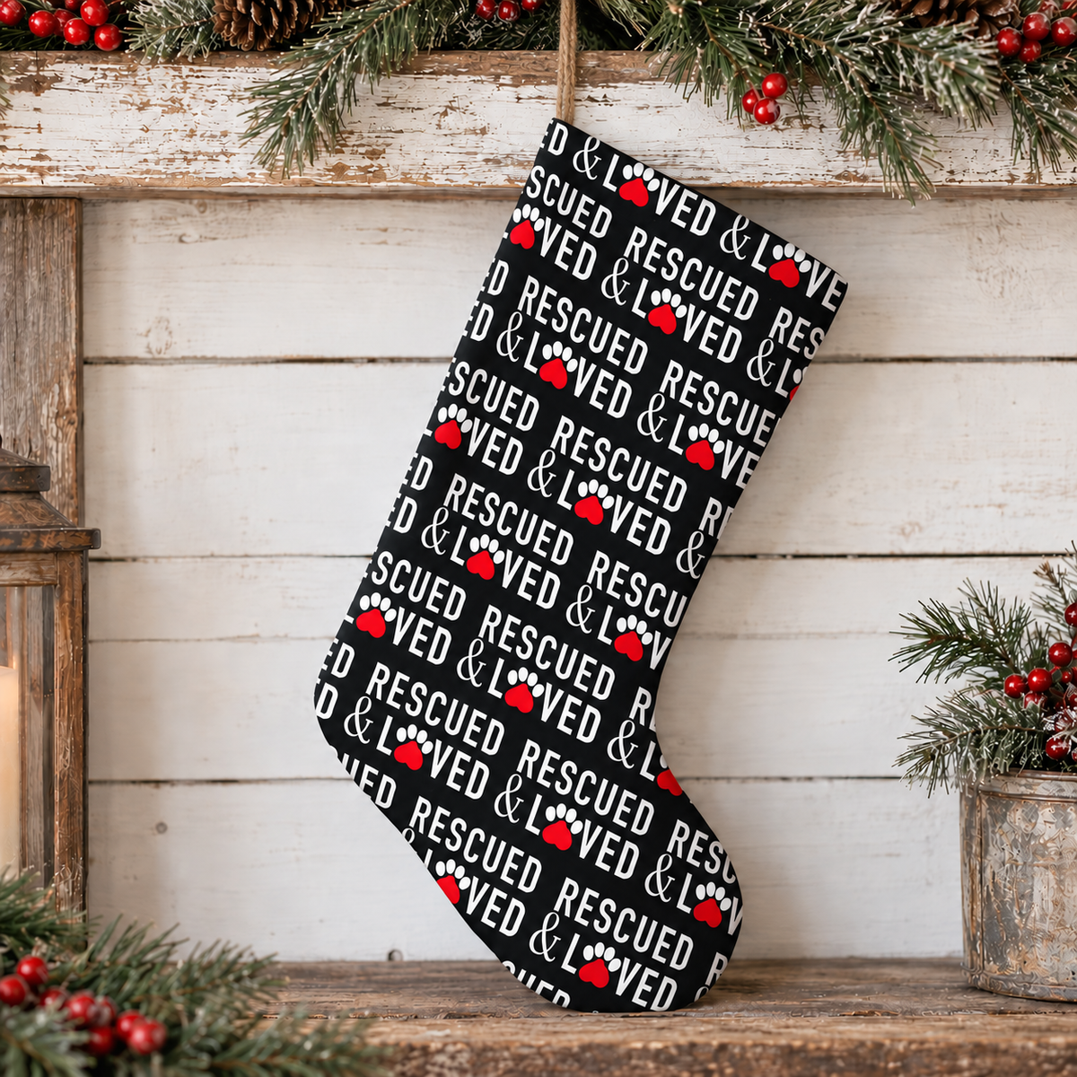 A Christmas stocking with a black background and a repeating pattern of white 'LOVED & RESCUED' text and red paw prints, hanging in front of a rustic white wood paneled wall decorated with pine branches and red berries.