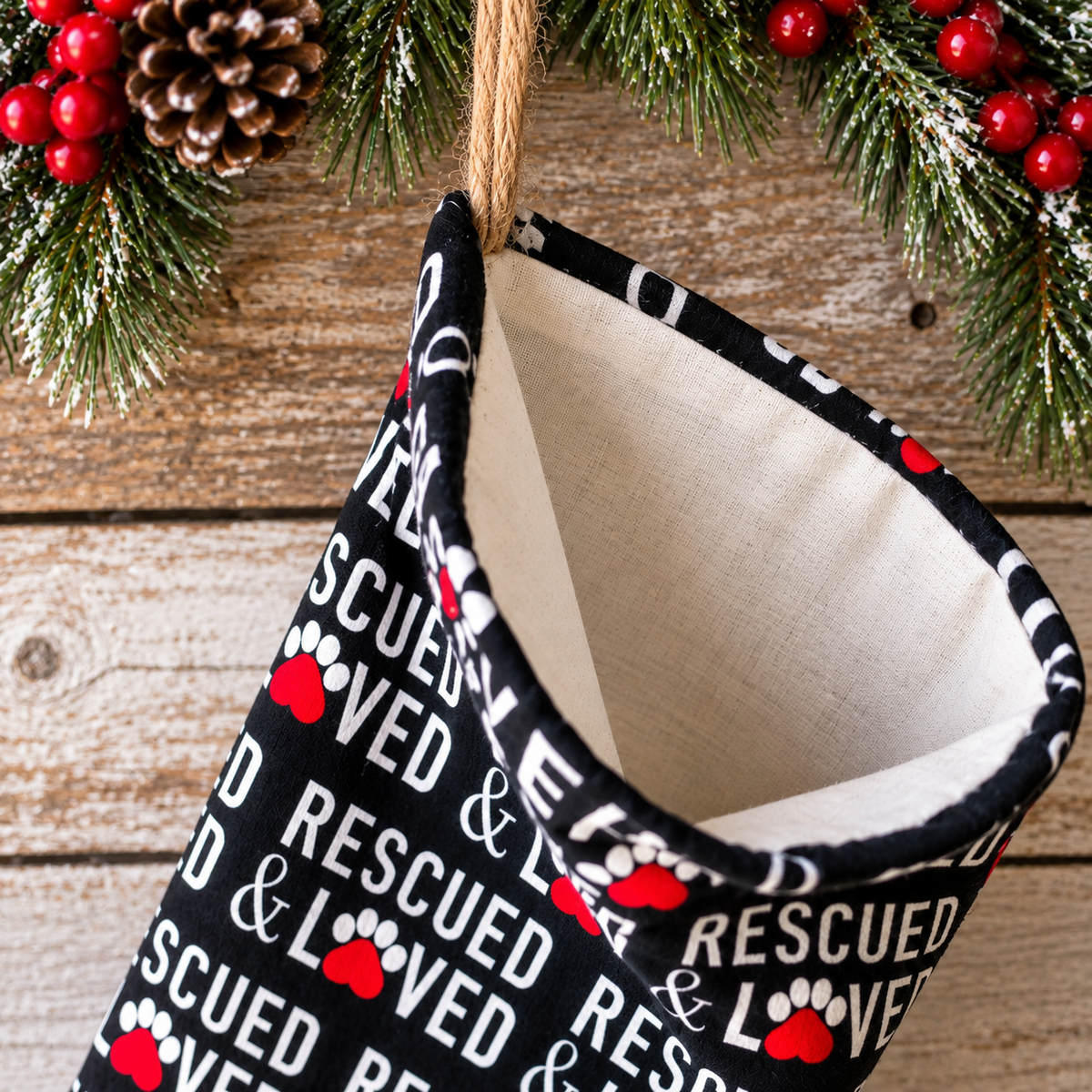A Christmas stocking with a black exterior printed with 'LOVED & RESCUED' in white text and red paw prints with hearts, hanging from a wooden surface with holiday greenery and pinecones.