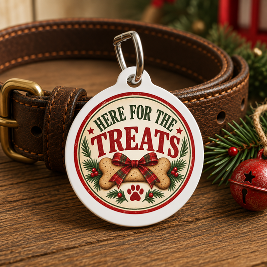 A round white pet ID tag with a silver clasp, attached to a brown leather collar. The tag features a festive Christmas design with the text 'HERE FOR THE TREATS' in red and green, a bone graphic with a red plaid bow, pine branches, and a red paw print, displayed on a wooden surface with holiday decorations.