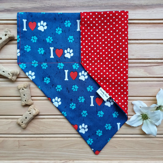 A reversible dog bandana with a 'I Love Dogs' print on one side and red polka dots on the other, displayed on a wooden surface with dog treats and a tag, with a white flower in the background.
