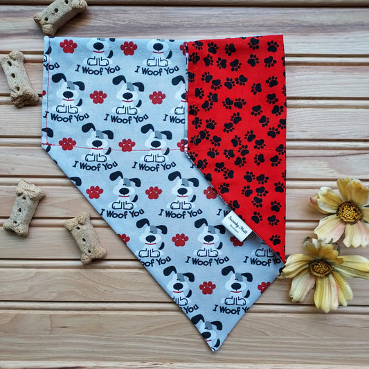 A reversible dog bandana with a 'I Woof You' text pattern on one side and a red paw print pattern on the other, displayed on a wooden surface with dog treats and flowers nearby.