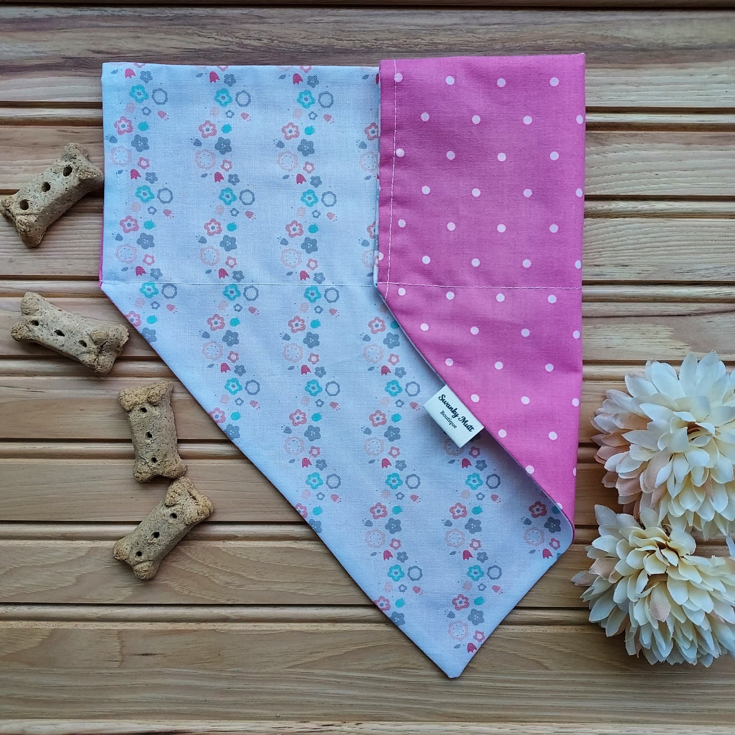 A reversible dog bandana with a pink flower and pink dot pattern on one side, and a similar pattern with green circles on the other, displayed on a wooden surface with cookies and a tag, with a pink flower accessory to the side.