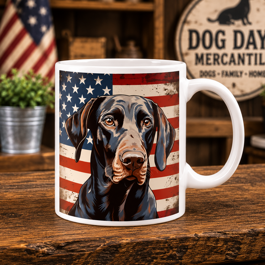 A white ceramic mug featuring a printed graphic of a German Shorthaired Pointer in front of a distressed American flag, set on a rustic wooden table with a blurred background containing a sign that reads 'DOG DAYS MERCANTILE'.
