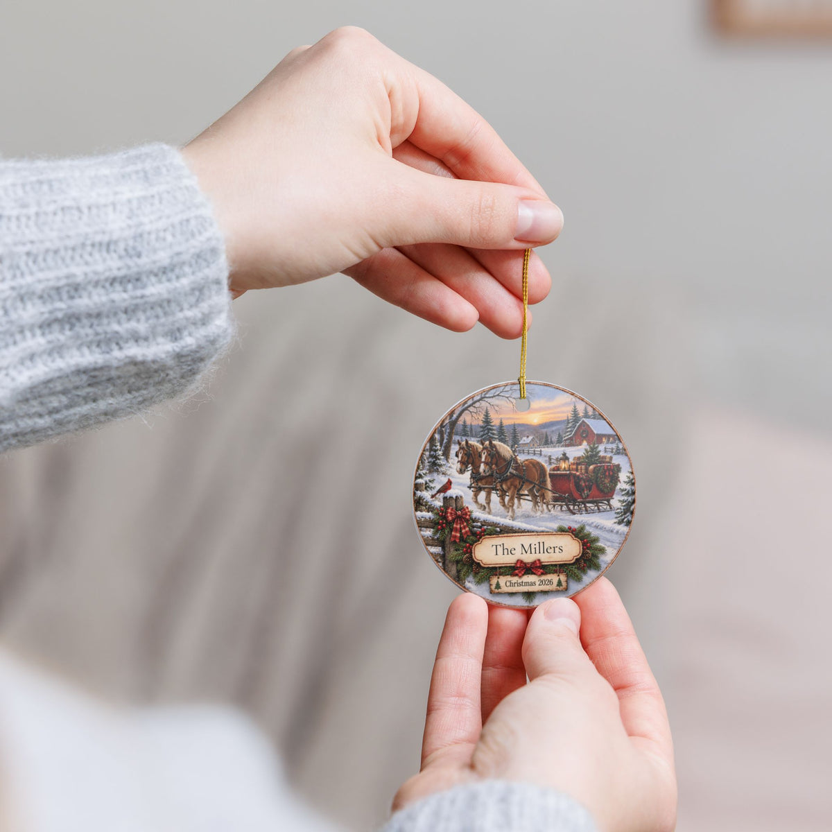 A person holds a round ceramic Christmas ornament featuring a winter scene with a horse-drawn sleigh and the text 'Name' and 'Winter 2022', shown against a soft, blurred indoor background.