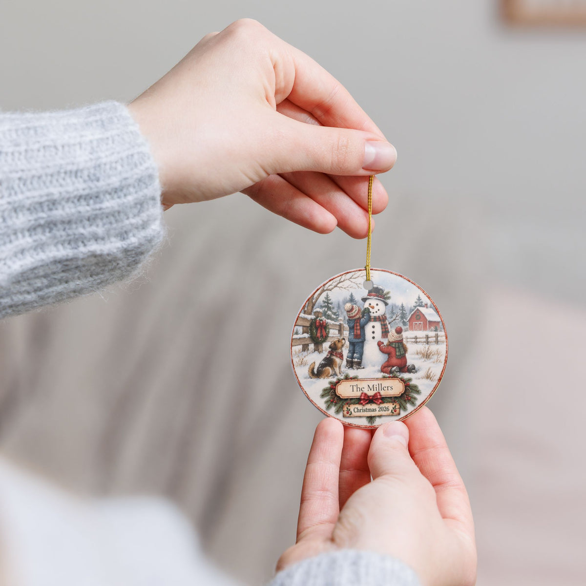 A person holds a round wooden Christmas ornament featuring a winter scene with a snowman.