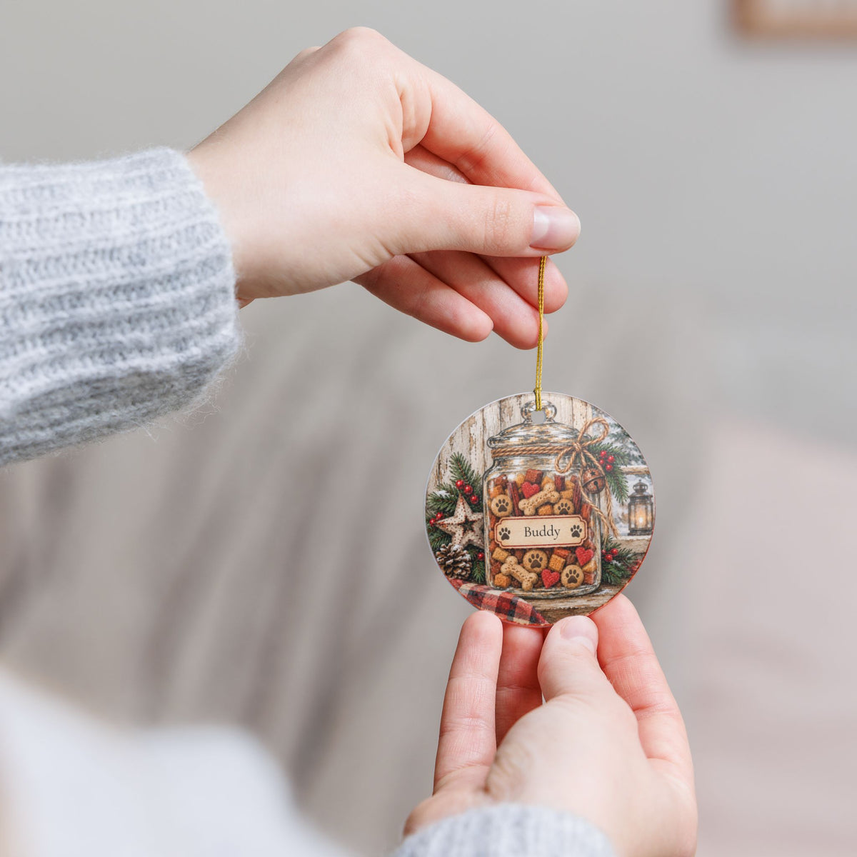 A person holds a round hanging ornament featuring a vintage-style illustration of a jar filled with cookies and the word 'Buddy.' The ornament is suspended by a thin gold string against a softly blurred indoor background.