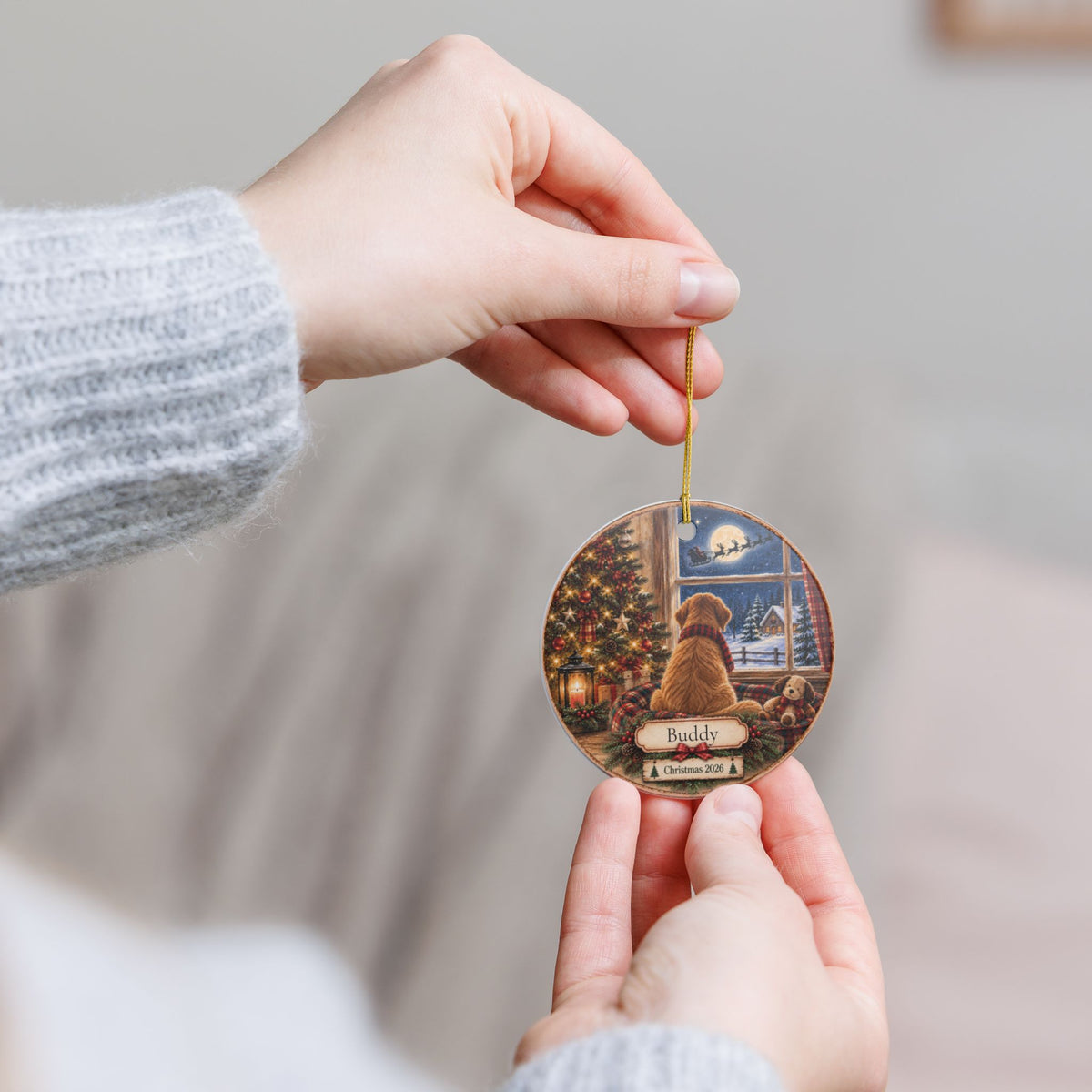 A person holds a round Christmas ornament featuring a dog sitting by a window next to a decorated Christmas tree. The ornament has a gold hanging string and is shown against a soft, blurred indoor background.