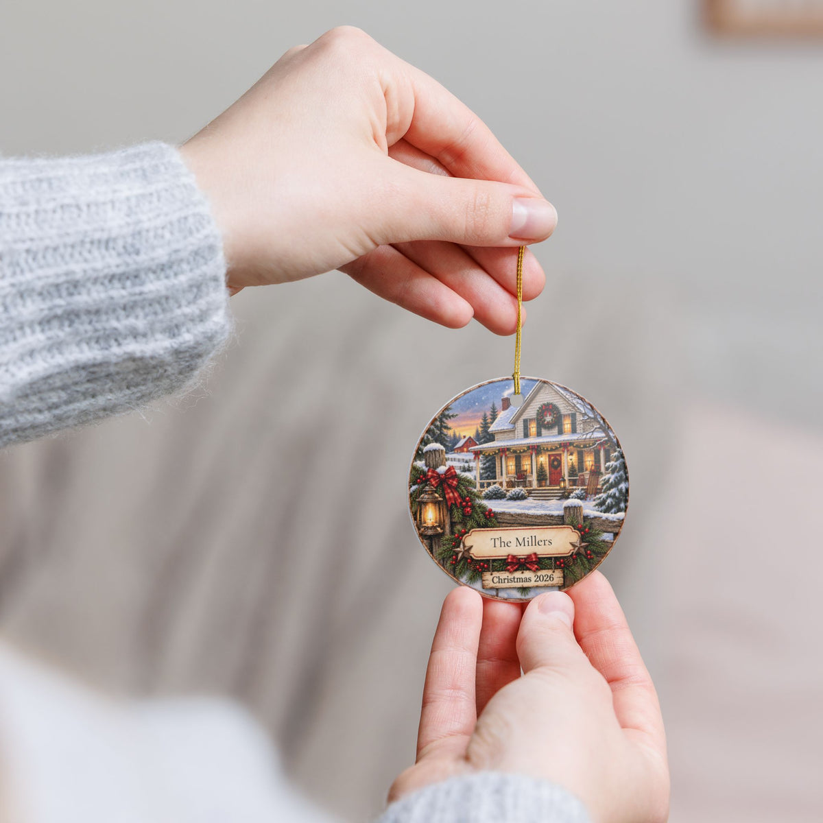 A person holds a round, personalized Christmas ornament featuring a snowy house scene with the text 'Christmas 2026' on a gold string, against a soft, blurred indoor background.