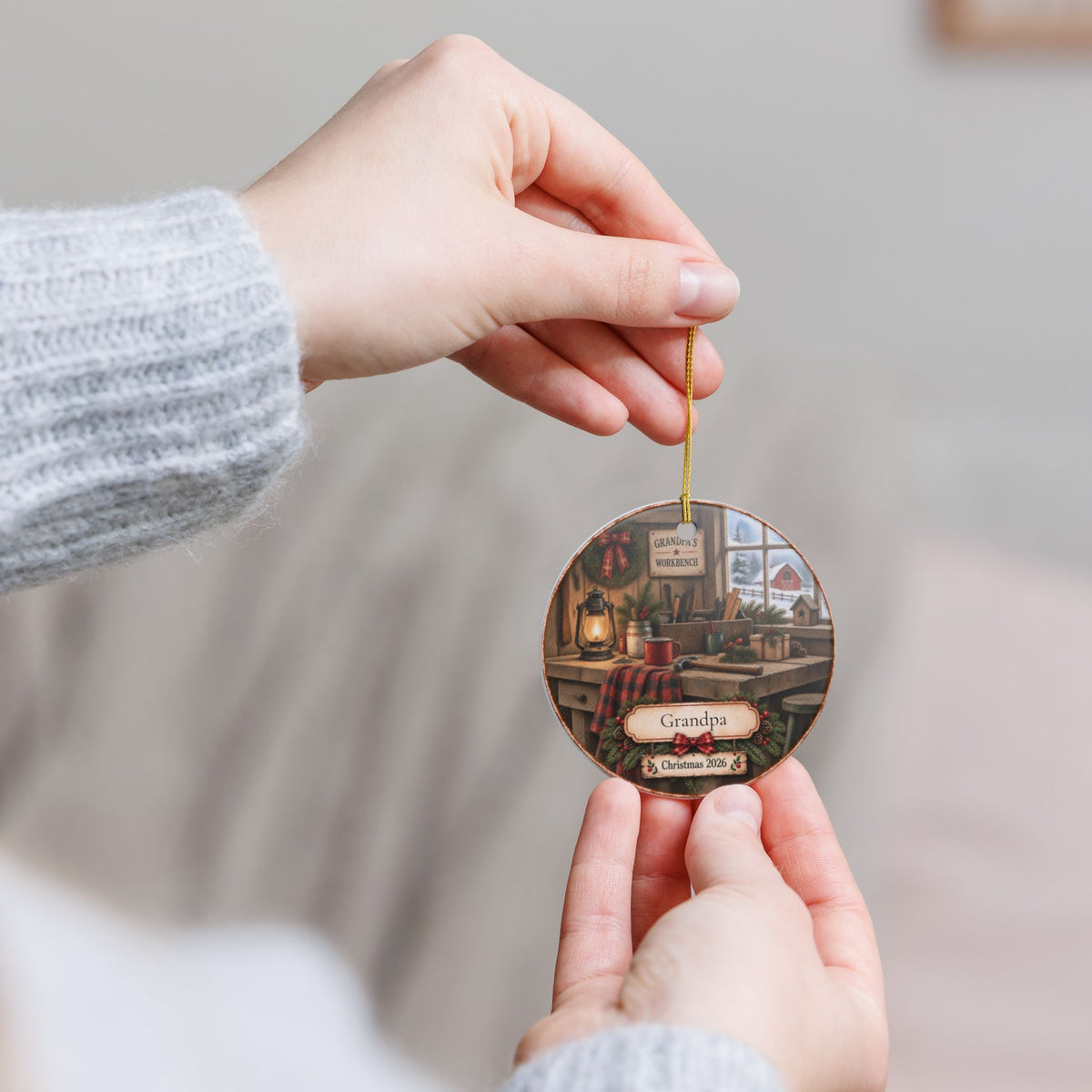 A person holds a round ceramic ornament featuring a cozy kitchen scene with the text 'Grandpa' and 'Christmas 2026' on a soft neutral background.