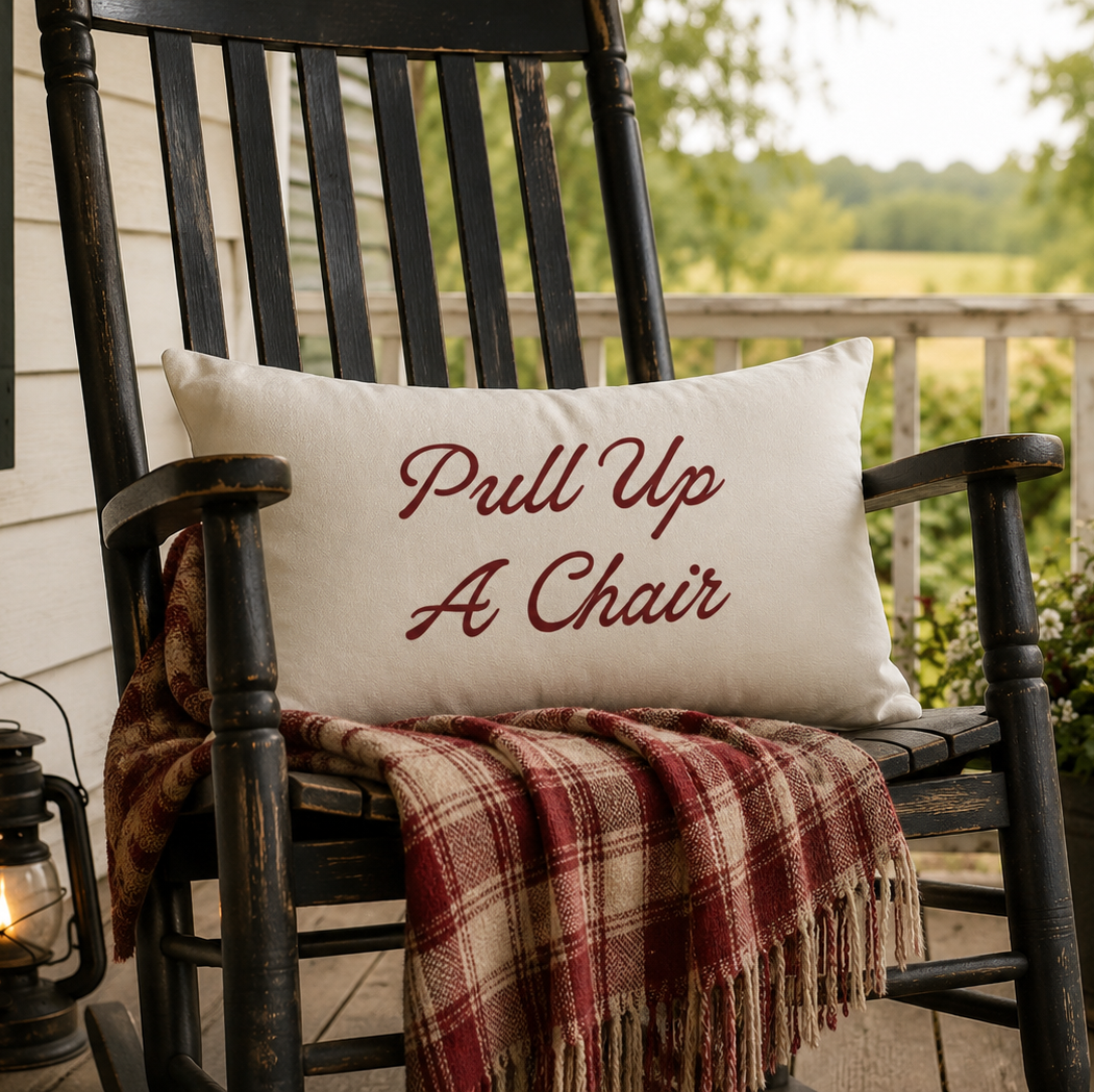 A white rectangular throw pillow with the phrase 'Pull Up A Chair' in red script, resting on a dark wooden rocking chair on a porch. A red and beige plaid blanket is draped over the chair, with a lantern on the left and a potted American flag and flowers on the right.