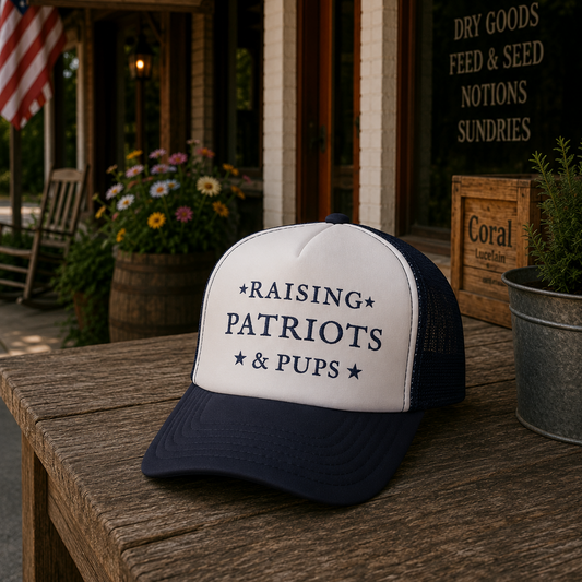 A navy blue and white trucker hat with the text '*RAISING PATRIOTS *PUPS *' on the front, displayed on a rustic wooden table outside a shop with a sign reading 'DOG DAYS MERCANTILE'.