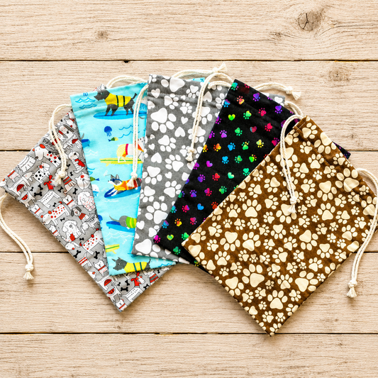 Fabric drawstring pouches with animal and pattern prints are arranged in a fan shape on a light wood plank background. The designs include a gray cartoon dog pattern, a light blue sea life pattern, a gray paw print pattern, a black multicolor paw print pattern, and a brown paw print pattern.