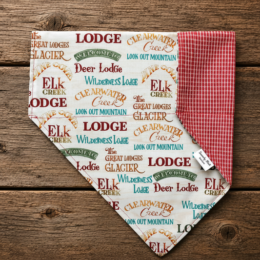 A triangular dog bandana laid flat on a rustic wooden surface. The front side has an allover print of lodge names and mountain phrases in red, green, and blue on an off-white background.
