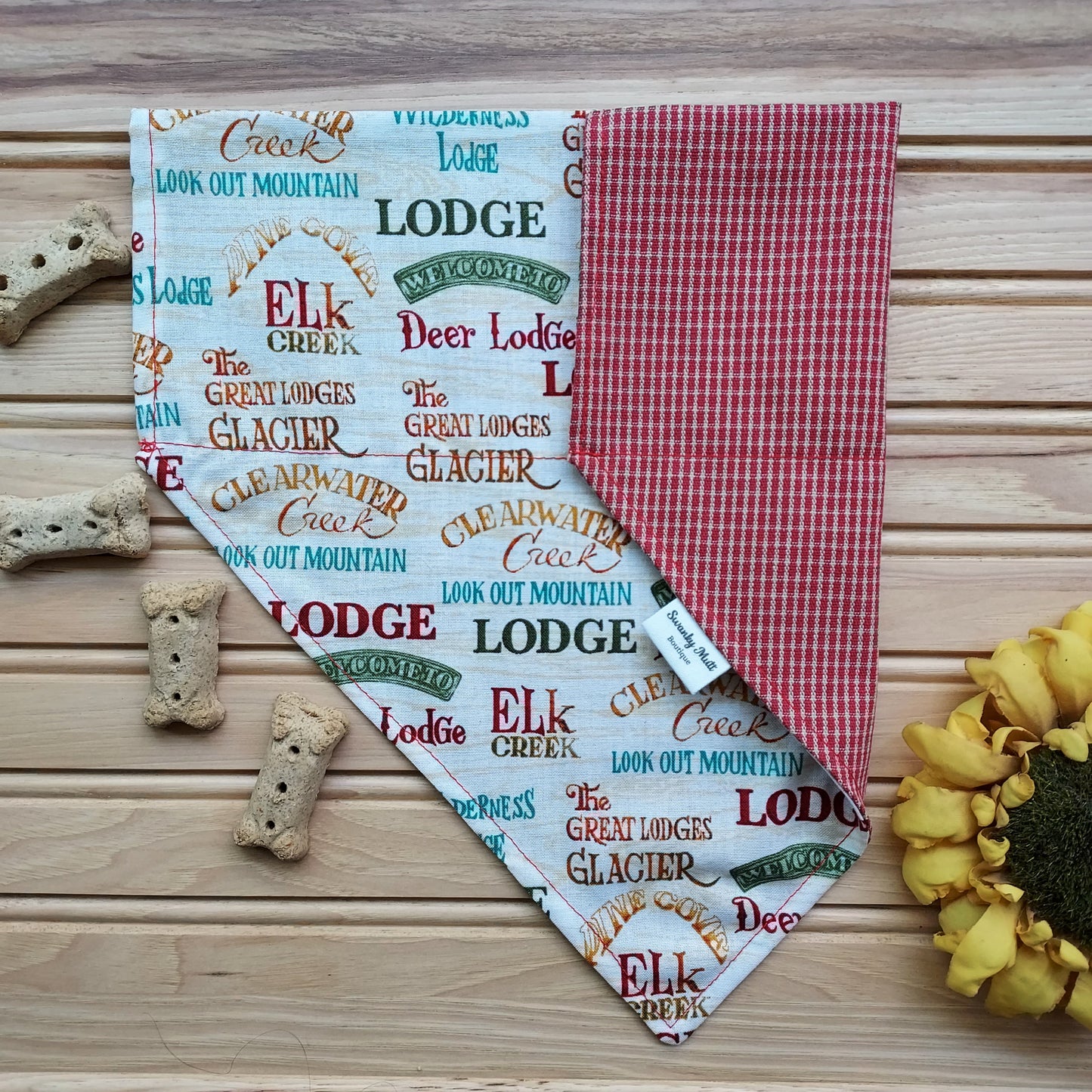 A reversible dog bandana with a rustic lodge print on one side and a rust plaid pattern on the other, displayed on a wooden surface with paw prints and dog treats around.