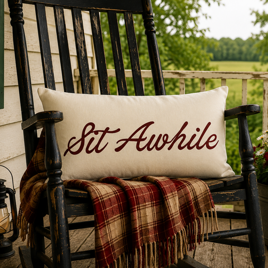 A cream rectangular throw pillow with the phrase 'Sit Awhile' in dark red script, displayed on a black wooden porch rocking chair. A red and beige plaid blanket is draped over the chair, with a lit lantern on the left and a potted American flag and flowers on the right, set against a background of a green field and trees.