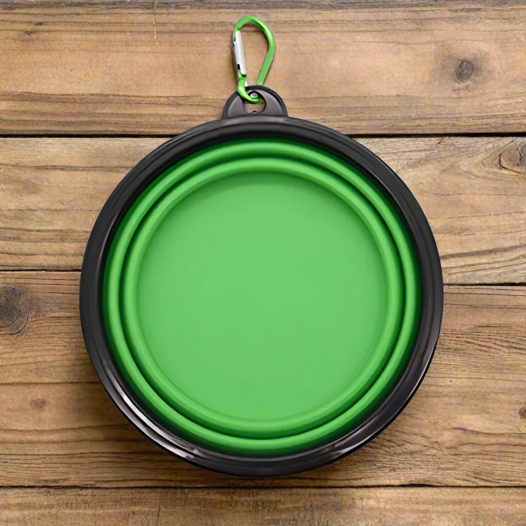 A green collapsible pet bowl with a black rim and a green carabiner clip, shown on a rustic wooden surface.