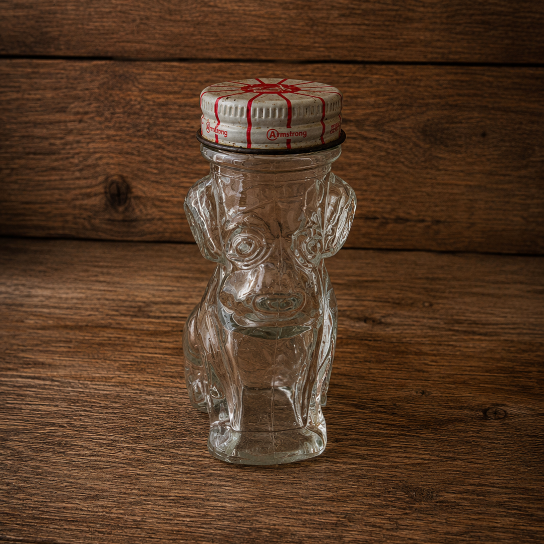 A small clear glass bottle shaped like a dog, with a white screw cap printed with a red design, standing on a rustic dark wood surface.